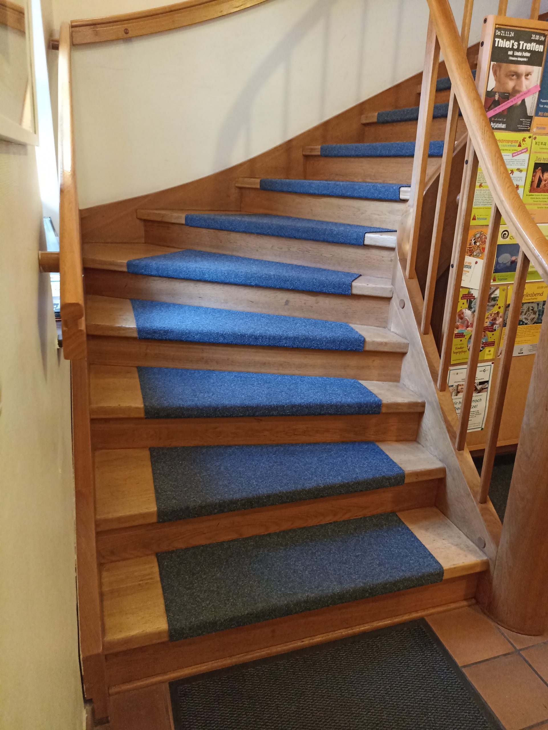 Das Bild zeigt eine Holztreppe mit Handlauf auf beiden Seiten. Die breiten Holzstufen sind mittig über ca. 70 cm mit grau-blauem Teppich belegt. Nach fünf Stufen verläuft die Treppe im Knick um neunzig Grad nach rechts.