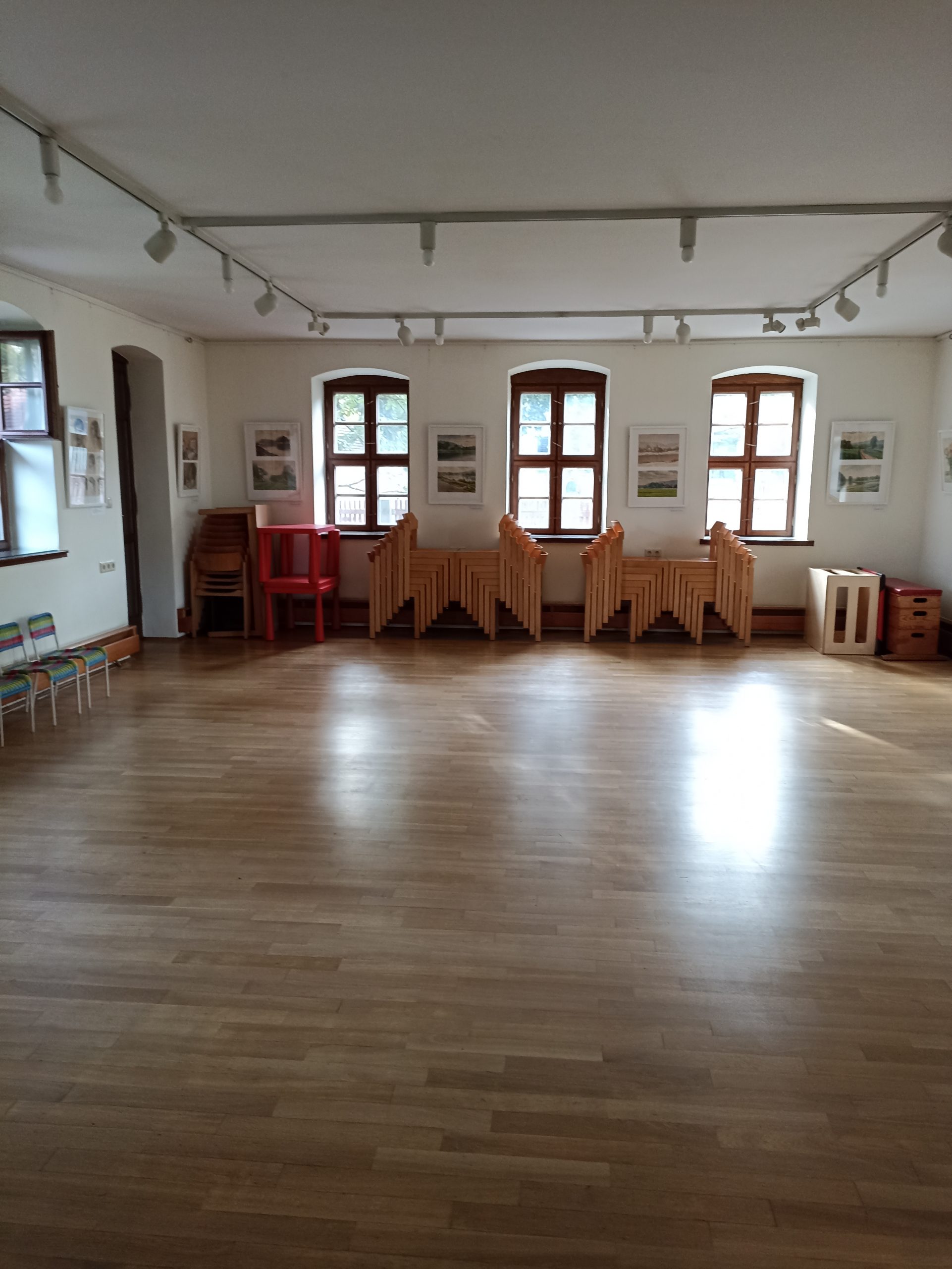 Auf dem Bild ist der untere Saal im Haupthaus zu sehen. Der sogenannte Kreativraum ist mit Parkett ausgekleidet. Das Bild zeigt den Blick von der Eingangstür in den Raum hinein auf die drei Südfenster im Hintergrund. Am hinteren Ende sind unter den Fenstern über die gesamte Länge gestapelte Stühle und Tische zu sehen. Zwischen den Fenstern hängen jeweils zwei Ausstellungsbilder.