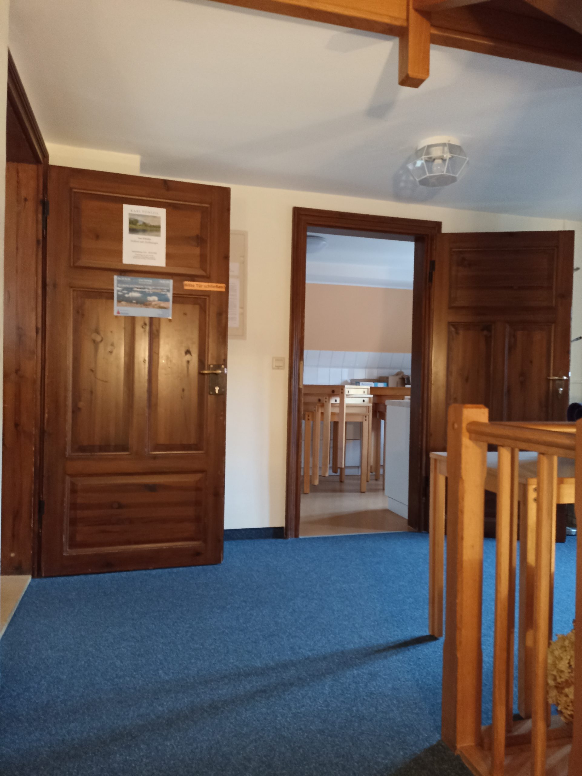 Das Bild zeigt den mit blauem Teppich ausgelegten Eingangsbereich des Obergeschosses im Haupthaus. Rechts ist im Vordergrund das Ende des hölzernen Handlauf der Treppe zu erkennen. Links ist die geöffnete Holztür zum Dachsaal und geradeaus die geöffnete Holztür zur Küche zu sehen.