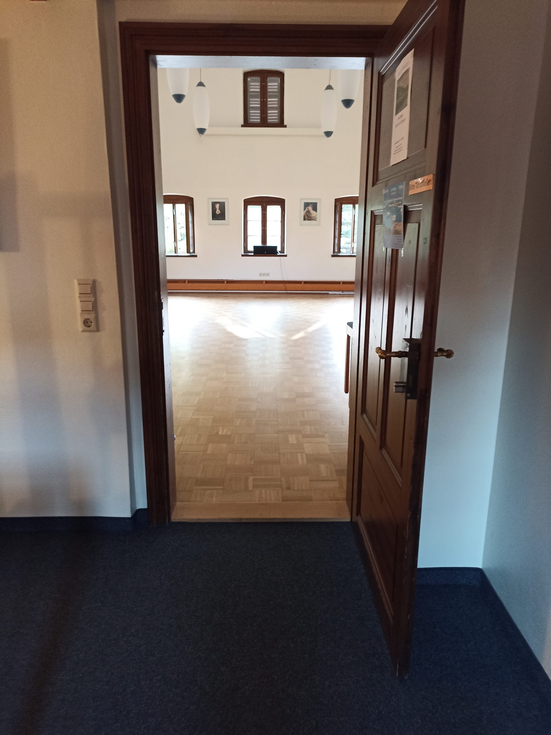 Das Bild zeigt den Blick aus dem Eingangsbereich des Obergeschosses durch die geöffnete Holztür in den Dachsaal. Bis zur zwei Zentimeter hohen Holzschwelle der Dachsaaltür ist der Boden mit blauem Teppich bedeckt, der Dachsaal selbst ist mit Parkettboden ausgelegt. Im Hintergrund sind die drei Fenster der Südseite zu sehen. Auf dem Parkett davor leuchtet das Sonnenlicht.